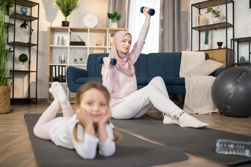 Wall Mural - Attractive positive happy muslim mom doing sports using dumbbells while her cute little girl is resting lying on the mat in living room at home.