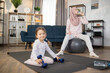 © sofiko14 - Resting after morning sports, young beautiful Muslim mother in a hijab sits on a fitball and drinks water while her little daughter sits on mat with dumbbells in her hands.
