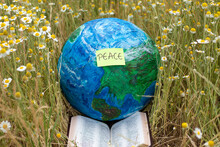 Earth Globe With A Handwritten Word Peace On A Note And Open Holy Bible Book In A Field With Green Grass And Flowers. Christian Biblical Concept Of 1000 Years (Millennium) Of Jesus Christ's Reign.