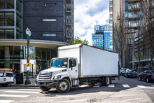 Big Rig Day Cab White Semi Truck With Long Box Trailer Making Local Commercial Delivery At Urban City With Multilevel Residential Apartments Buildings Turning On The City Street With Crossroad