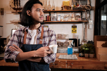 Wall Mural - Long hair handsome Asian male startup barista with an apron stands at a casual cafe, arms crossed, looking outside with a coffee cup, alcohol sanitizer on the counter bar, coffee shop service jobs.