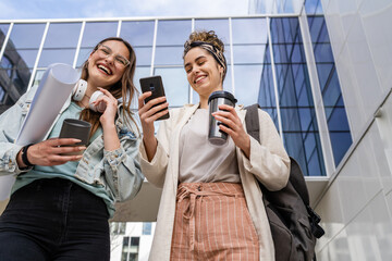 two women female students walk in front of modern university building with notes and paper documents using mobile phone discuss talk happy smile while holding mobile phone reading sms text messages