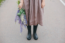 Cottagecore Aesthetics. Close Up Of Woman In Rustic Dress And Gumboots Holding Lupine Bouquet In Summer Countryside. Young Female With Wildflowers After Rain On Rural Road. Moody Image