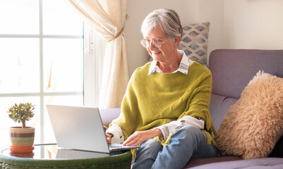 Smiling elderly woman working on laptop at home sitting on sofa in living room. Modern senior lady with green poncho browsing internet
