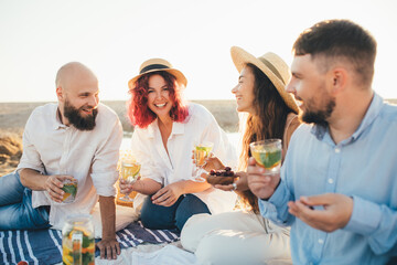 Group of happy friends having picnic, drinking lemonade and eating fruits, sitting near sea at sunset.