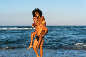 Happy young interracial couple in piggybak on beach – man giving piggybak his woman and laughing at tropical beach – smiling boyfriend in love carrying on back her girlfriend and having fun 