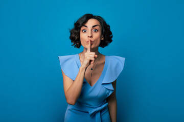 Wall Mural - Close-up photo of a beautiful girl in a blue dress, who is looking in the camera while pressing her index finger to her lips and isolated on blue background