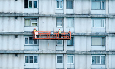 Two plasterers-painters are painting facade of house, standing in construction cradle. View of facade from the back of workers, restoration process at height in lifting bucket