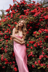 Wall Mural - beautiful woman with blond hair in elegant pink dress posing in blooming garden with red roses