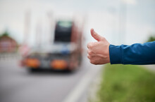 Close Up Of Man's Hand Hitchhiking By Roadside. Male Hand Showing Thumbs Up Gesture Outdoors On Blurred Background. Hitchhiking, Hitching, Auto Stop Concept.