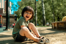 A Three-year-old Asian Boy With Long Black Hair Is Sitting On The Sand And Wearing Sandals. A Child Walks On A Hot Summer Day In A City Park. Barefoot Boy