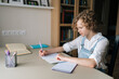 © dikushin - Medium shot of little child schoolgirl writing in exercise book doing some homework, sitting at table near window. Cute curly small school girl studying alone making notes at home.