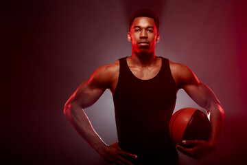 Wall Mural - Basketball player side lit with red color holding a ball against smoke background. Serious concentrated african american man