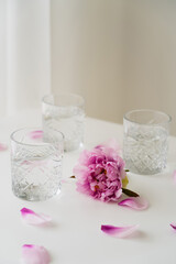 Sticker - pink peony and floral petals near glasses with fresh water on grey background.