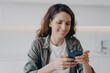 © VK Studio - Spanish girl in casual wear is typing and receiving messages. Happy woman with smartphone at home.