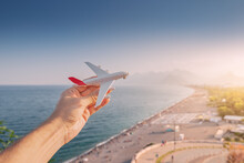 Toy Airplane In Hand Against The Background Of The Famous Konyaalti Beach In Antalya - The Main Turkish Resort And Riviera. Passenger Traffic And Flights To Dream Vacation