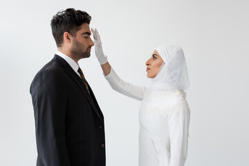 Wall Mural - side view of young bride in hijab touching forehead of groom during wedding ceremony isolated on grey.