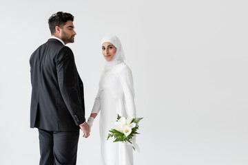 Wall Mural - smiling muslim bride in wedding dress with bouquet of calla lily holding hands with groom isolated on grey.