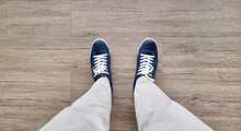 Feet In Denim Sneakers Standing On Wooden Vinyl Floor, Selective Focus. Fashion Hipster Cool Man With Wblue Sneakers And Beige Pants, Soft Vintage Toned Colors For Beginning Concept.