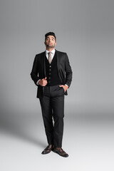 Wall Mural - full length of stylish muslim groom in suit posing with hand in pocket while standing on grey.