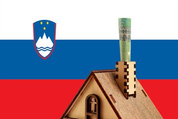 Euro bill in a chimney of a wooden toy symbolic house against the background of the Slovenia flag