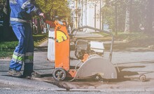 A Road Worker In Overalls Uses A Portable Asphalt Cutter To Cut Asphalt With A Diamond Blade To Repair Part Of The Roadway. Cutting Road Works With A Gasoline-powered Angle Grinder. Road Repairs.