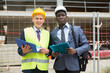 African-american architect and a young engineer controlling the progress of work are standing on a construction site, holding a laptop and an estimate in their hands
