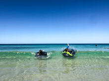 Young Boys In Wetsuits Lying On Inflatable Tyres On Small Beach Waves