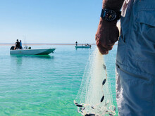 Close Up Of Mans Hand Holding Fishing Throw Net On Clear Aqua Ocean With Two Dinghies In Background