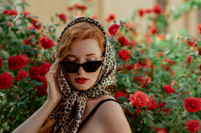 Beautiful Fashionable Redhead Freckled Woman Wearing Trendy Black Cat Eye Sunglasses, Leopard Print Headscarf, Posing In Red Rose Garden. Copy, Empty Space For Text