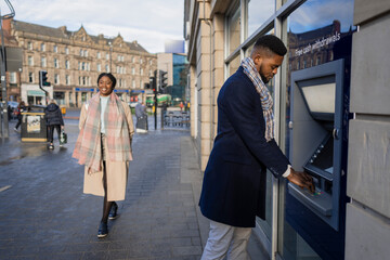 Wall Mural - Man Using Cash Machine With Woman Walking on Pavement