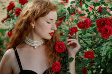 Beautiful Redhead Freckled Woman Wearing Elegant Pearl Necklace, Golden Earrings, Posing In Red Rose Garden. Copy, Empty Space For Text
