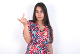 young beautiful brunette woman wearing colourful dress over white wall shows middle finger bad sign asks not to bother. Provocation and rude attitude.