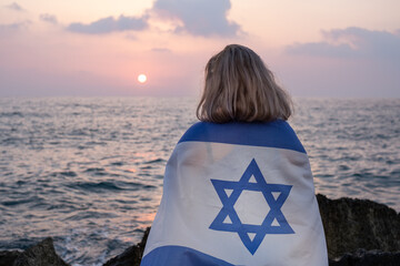 Teen female with Flag of Israel draped over her shoulders. Patriotic israeli young woman enjoy sunset on sea