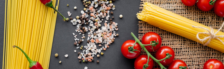 Poster - Top view of pasta near spices and vegetables on sackcloth on black background, banner.