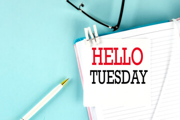 HELLO TUESDAY text on a sticky on notebook with pen and glasses , blue background