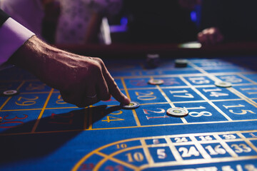 Vibrant casino table with roulette in motion, with casino chips, tokens, the hand of croupier, dollar bill money and a group of gambling rich wealthy people playing bet in the background