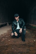 © Alexander White - young male model in black leather jacket and cap kneeling down in a tunnel with chelsea boots