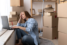 Young Worried Unhappy Woman Sitting On The Floor Near Moving Boxes Using A Laptop With Hands On Head Suffering Of Headache, Searching For A New Apartment