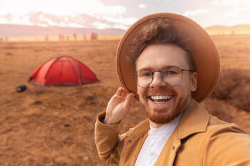 Wall Mural - Happy hiker man with hat making selfie photo portrait background red tent mountain. Concept travel blogger, adventure life trip