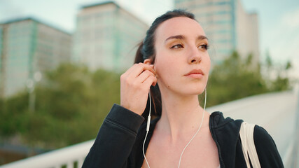 Young woman with freckles and long ponytail wearing black sports hoodie in wired headphones listens to music and walks along the bridge