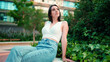 © Andrii Nekrasov - Young woman with freckles and dark loose hair wearing white top and jeans is enjoying the view of modern city. Girl with long eyelashes sits in public garden on modern building background
