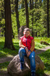 © Alina Lebed - A young man in a sleeveless red jacket is sitting on a huge stone in a pine forest in spring. The magical light from the sun's rays falls behind the boy.