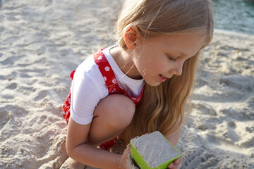 A pretty little girl with blonde hair 5 years old is playing outside in the sand with toys