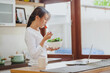 © interstid - Happy Asian woman eating healthy salad while workinh with laptop on table in kitchen at home