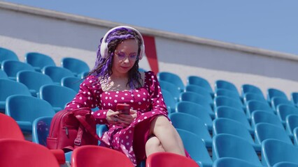 Wall Mural - Young relaxed woman in glasses, headphones sitting on stadium bleachers alone, listening to music, dancing outdoors in summertime. Alone lady in red dress relaxing at street stadium, listening to song