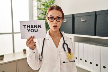 Poster - Young caucasian woman wearing doctor uniform holding thank you banner scared and amazed with open mouth for surprise, disbelief face