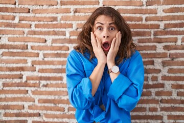 Wall Mural - Beautiful brunette woman standing over bricks wall afraid and shocked, surprise and amazed expression with hands on face