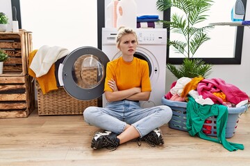 Poster - Young blonde woman doing laundry sitting by washing machine skeptic and nervous, disapproving expression on face with crossed arms. negative person.