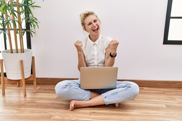 Canvas Print - Young blonde woman using computer laptop sitting on the floor at the living room very happy and excited doing winner gesture with arms raised, smiling and screaming for success. celebration concept.